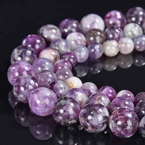 May include: A selection of spherical beads in varying sizes and shades of purple, lavender, and white. The beads have a polished, reflective surface. The arrangement suggests they are for jewellery making.