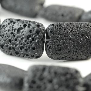 Puede incluir: Una hebra de cuentas de roca volcánica negra, cada cuenta es rectangular y tiene una textura rugosa y porosa.