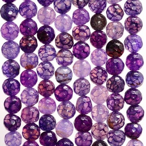 Könnte beinhalten: Eine Strähne aus violetten Achatperlen mit einer rissigen, strukturierten Oberfläche. Die Perlen sind rund und haben eine hellviolette Farbe mit dunkleren violetten Adern.