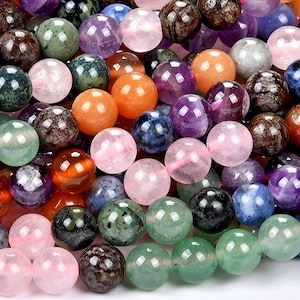 May include: A close-up of a variety of round gemstone beads in various colors, including pink, green, purple, blue, and brown. The beads are smooth and polished, and they are arranged in a random pattern.