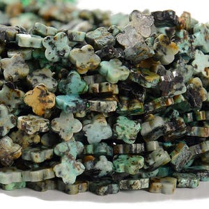 May include: Close-up of a pile of turquoise-colored gemstone beads. The beads are shaped like four-leaf clovers, with variations in color including shades of green, brown, and blue. The beads are likely used for jewelry making or crafting.