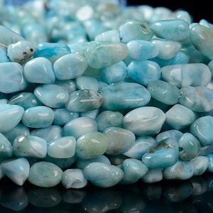 May include: A close-up of a collection of tumbled, polished gemstones. The stones are primarily light blue with hints of white and green. The smooth, rounded stones are strung together, suggesting they are beads for jewelry making.