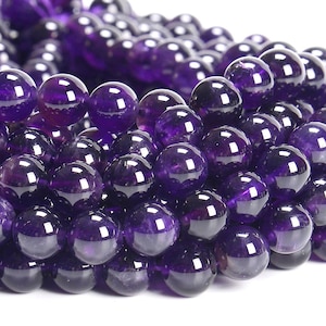 May include: Close-up of a collection of polished, round amethyst beads. The beads are a deep, rich purple color with a glossy finish, reflecting light. The beads are strung together, suggesting they are for jewelry making or crafting.