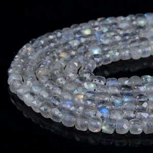 May include: A strand of white faceted gemstone beads with iridescent blue and green hues. The beads are square shaped and have a smooth, polished finish.