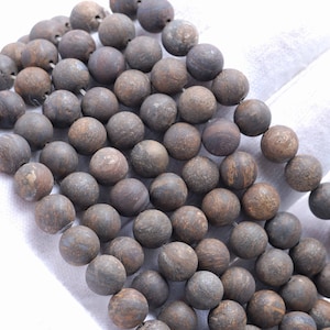 Puede incluir: Una hebra de cuentas redondas de color marrón oscuro y gris hechas de piedra natural. Las cuentas tienen una superficie rugosa y texturizada.