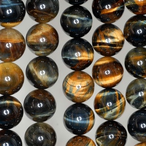 Puede incluir: Primer plano de múltiples cuentas pulidas de piedra ojo de tigre. Las cuentas son esféricas y muestran una mezcla de colores marrón dorado, marrón oscuro y azul grisáceo. Las cuentas tienen una superficie reflectante y brillante.