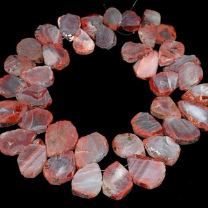 Op de afbeelding: Een streng rode agaat edelsteen kralen, elke kraal is een unieke, onregelmatige vorm met een natuurlijke, ruwe rand. De kralen zijn geregen op een draad en hebben een doorschijnende, rood-witte kleur.