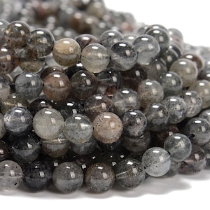 May include: A close-up of a pile of round beads. The beads are a mix of gray, brown, and clear colors, with a speckled appearance. The beads are smooth and shiny, and appear to be made of a natural stone material. The beads are strung together.