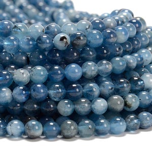 May include: A strand of round, blue gemstone beads with a slightly cloudy appearance. The beads are smooth and polished, and they have a light blue color with a hint of gray. The beads are arranged in a random pattern, and they are all about the same size.
