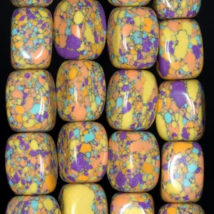 Puede incluir: Primer plano de una colección de cuentas rectangulares coloridas. Las cuentas presentan un patrón moteado en tonos de amarillo, naranja, morado y turquesa. Están dispuestas en filas ordenadas sobre un fondo negro, mostrando sus colores vibrantes y diseño único.