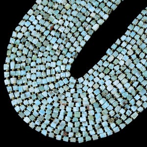 Puede incluir: Un collar de múltiples hilos compuesto por numerosas piedras de Larimar azul claro cortadas en forma cuadrada. Cada piedra presenta un veteado único en blanco y verde pálido, separadas por pequeñas cuentas metálicas doradas en arco.