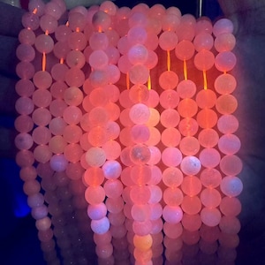 Puede incluir: Una hebra de cuentas de piedras preciosas rosadas y blancas, que brillan bajo una luz UV, con un tono naranja brillante. Las cuentas son redondas y lisas, y la hebra está enhebrada en un cordón elástico transparente.