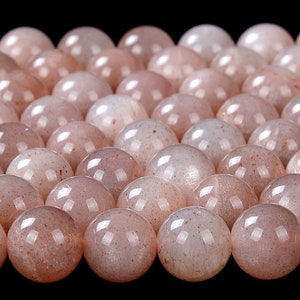May include: A close-up of a group of round, translucent, light pink sunstone beads. The beads have a subtle sparkle and a slightly iridescent sheen.