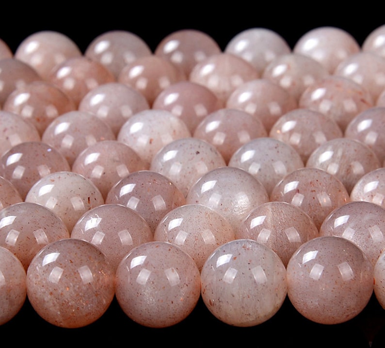 May include: A close-up of a group of round, translucent, light pink sunstone beads. The beads have a subtle sparkle and a slightly iridescent sheen.
