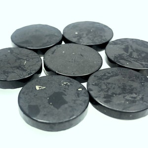 Puede incluir: Siete piedras redondas lisas de color negro con una textura ligeramente rugosa. Las piedras están dispuestas en círculo sobre un fondo blanco.