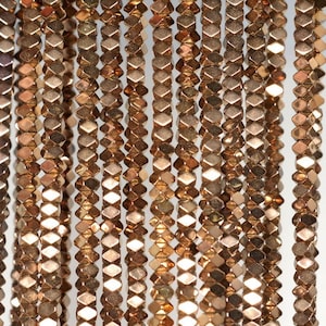 Op de afbeelding: Close-up van meerdere strengen metallic bronskleurige kralen. Elke kraal heeft een facetvormige, geometrische vorm die licht reflecteert. De kralen zijn aan elkaar geregen en creëren verticale lijnen tegen een witte achtergrond. Ideaal voor het maken van sieraden.