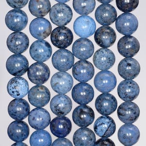 Op de afbeelding: Een streng blauwe edelsteen kralen. De bolvormige kralen variëren in blauwtinten, sommige vertonen donkere spikkels. Ze zijn geregen en vormen een doorlopende lijn van gepolijste bollen. Elke kraal heeft een diameter van ongeveer 1 cm.