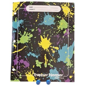 May include: A black Trapper Keeper portfolio featuring a vibrant paint splatter pattern in yellow, teal, and purple. The cover includes a white rectangular label area with the printed text NAME and SUBJECT, and three punched holes along the left side.