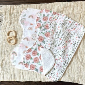 May include: Three baby cloth nappies with floral and rainbow patterns, plus a wooden teething ring. The nappies feature designs with pink roses, rainbows, and botanical elements. The items are arranged on a cream-coloured, textured surface.