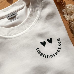 Op de afbeelding: Een wit sweatshirt met ronde hals en een zwarte opdruk op de rechterbovenkant van de borst. Het ontwerp bestaat uit twee kleine hartjes boven de tekst "Lieblingsmensch", geschreven in een gebogen, vetgedrukt, schreefloos lettertype.