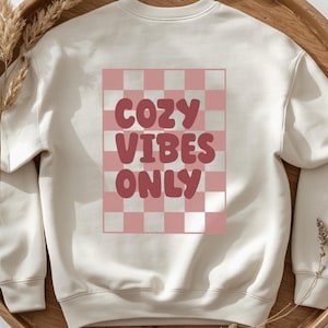 May include: A cream-colored crewneck sweatshirt featuring a rectangular graphic on the back. The graphic consists of a pink and white checkerboard pattern with the text COZY VIBES ONLY written in a bold, rounded, dark pink font across the center.