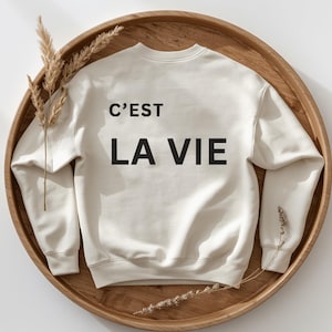 Könnte beinhalten: Ein cremefarbenes Sweatshirt mit Rundhalsausschnitt und dem schwarzen Schriftzug C'EST LA VIE auf der Vorderseite. Das Kleidungsstück hat lange Ärmel mit gerippten Bündchen sowie einen gerippten Saum und zeigt ein schlichtes Design auf hellem Stoff.