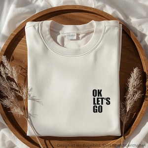 Puede incluir: Una sudadera de cuello redondo color crema con el texto negro "OK LET'S GO" impreso en la parte inferior derecha. La prenda tiene un cuello acanalado y una pequeña etiqueta de marca blanca en el interior del cuello, sobre una bandeja.