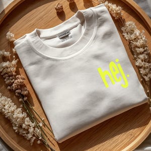 May include: A cream-colored sweatshirt with the word "hej" in bright yellow lettering. The sweatshirt is neatly folded on a wooden tray, surrounded by dried flowers. The overall aesthetic is minimalist and modern.
