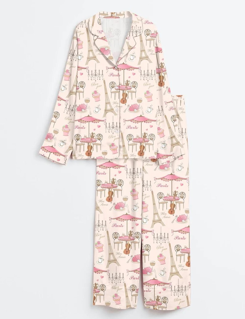 May include: Light pink two-piece pyjama set featuring a repeating pattern of Parisian cafe scenes, Eiffel Towers, and the word "Paris". The long-sleeved top has a button-down front, and the trousers are full-length.