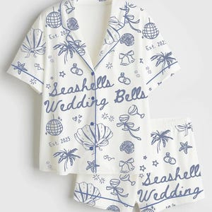 May include: White pajama set with blue seashell, wedding bell, and palm tree illustrations. The short-sleeved top and shorts feature the text "Seashells Wedding Bells" and "Est. 2025".