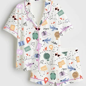 May include: White pajama set with a travel-themed print. The short-sleeved top and shorts feature illustrations of airplanes, cameras, globes, suitcases, and hearts. The set has pink trim and the text "XOXO".