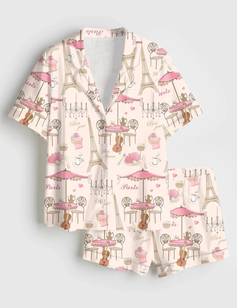 May include: Pale pink pajama set with a repeating design of the Eiffel Tower, cafe tables with umbrellas, violins, and cupcakes. The words "Paris", "Bon jour", and "Mon amour" are visible. The set comprises a button-down shirt and shorts.