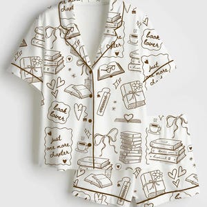 May include: White pajama set with a brown book-themed pattern. The set includes a short-sleeved button-up shirt and shorts, both adorned with book illustrations, hearts, and the text "book lover" and "just one more chapter."