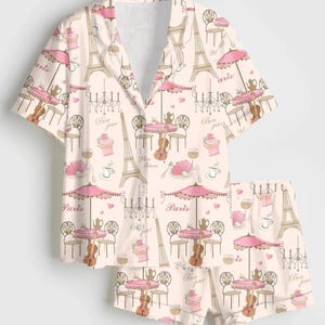 May include: Pale pink pajama set with a repeating pattern of the Eiffel Tower, cafe tables, violins, and cupcakes. The words "Paris", "Bon jour", and "Mon amour" are printed on the set. The set includes a short-sleeved shirt and shorts.