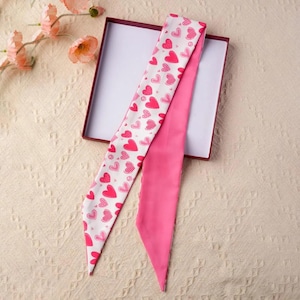 May include: A pink and white scarf with a heart pattern, and a solid pink scarf, are displayed inside an open maroon box. The patterned scarf has pink hearts on a white background. The solid pink scarf provides a contrasting colour.