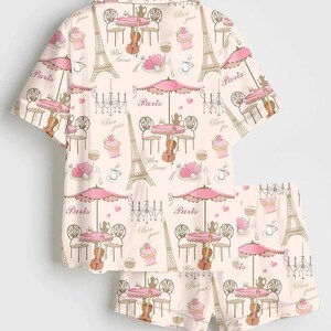 May include: A two-piece pyjama set with a repeating pattern of Parisian cafe scenes. The design includes the Eiffel Tower, cafe tables with umbrellas, violins, and the word "Paris" in a soft pink and beige colour scheme.