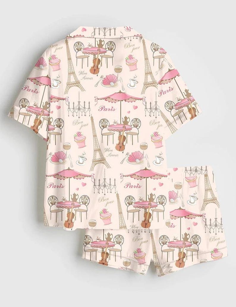 May include: A two-piece pyjama set with a repeating pattern of Parisian cafe scenes. The design includes the Eiffel Tower, cafe tables with umbrellas, violins, and the word "Paris" in a soft pink and beige colour scheme.