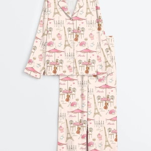 May include: A light pink pajama set featuring a repeating pattern of Parisian cafe scenes, including the Eiffel Tower, tables with umbrellas, and musical instruments. The set includes a button-down top and matching trousers.