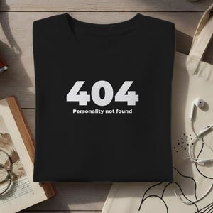 Könnte beinhalten: Schwarzes T-Shirt mit dem weißen Text "404 Personality not found". Das Shirt wird auf einer Holzoberfläche zusammen mit einer Vintage-Kamera, einem offenen Buch und einer Tragetasche mit weißen Ohrhörern präsentiert.