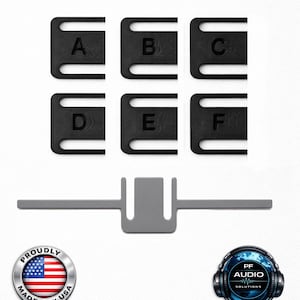 Puede incluir: Seis clips rectangulares negros etiquetados de la A a la F, cada uno con dos ranuras paralelas. Debajo hay un solo clip gris en forma de T. La imagen incluye una insignia circular Proudly Made in USA, un logotipo de PF Audio Solutions, un logotipo de UltiMaker y un logotipo de Atomic Filament.