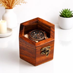 May include: A hexagonal wooden watch box with a dark brown finish and a brass-coloured metal latch. Inside the open box sits a round wristwatch featuring a black dial with white markers, silver hands, and three sub-dials on a light-coloured cushion.