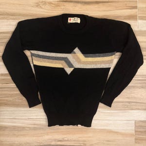 May include: A black long-sleeve sweater featuring a horizontal geometric pattern across the chest in shades of grey, beige, and white. The garment has a crew neckline, ribbed cuffs, and a hem, with a label at the neck reading Pringle of Scotland.