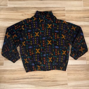 Könnte beinhalten: Eine schwarze Fleecejacke mit durchgehendem Frontreißverschluss, Stehkragen und langen Ärmeln. Der Stoff ist mit einem geometrischen Muster aus orangefarbenen X-Formen, violetten Winkeln sowie kleinen türkisfarbenen und roten Strichen bedruckt.