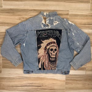 May include: A light blue denim jacket featuring a distressed collar and white paint splatters across the shoulders and sleeves. The back panel displays a large graphic print of a skull wearing a feathered headdress with the text YEEZUS above it.