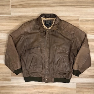 May include: A brown leather bomber jacket featuring a fold-down collar, a front zipper closure, and two large zippered chest pockets. The jacket includes dark olive green ribbed cuffs and a matching ribbed hem, with a light-colored interior lining.