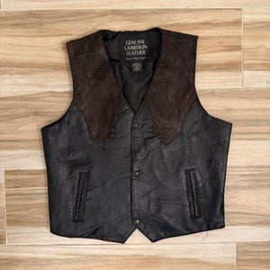 May include: A black leather vest featuring dark brown snakeskin-textured leather panels on the upper chest. The vest has a V-neckline, three front snap buttons, and two vertical slit pockets. The interior label reads GENUINE LAMBSKIN LEATHER.