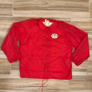 May include: A bright red nylon coach jacket featuring a classic collar, long sleeves with elastic cuffs, and a front snap-button closure. An oval patch on the left chest displays the text "TOMATOES OF RUSKIN" with a small tomato graphic.