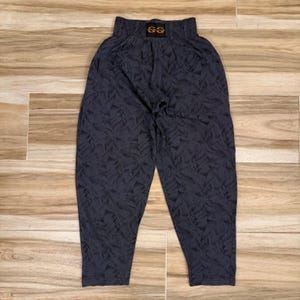 May include: Dark gray pants featuring an all-over abstract geometric pattern of angled lines and shapes. The pants have an elasticized waistband with a black rectangular label at the center front displaying the orange text GG.