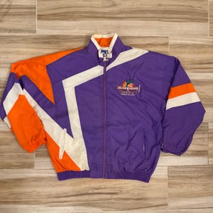 Puede incluir: Una chaqueta cortavientos con cremallera en color morado, naranja y blanco, con cuello alto y mangas largas. El pecho muestra un logotipo bordado con el texto All Star Weekend Phoenix February 10-12, 1995. Tiene una cremallera frontal completa.