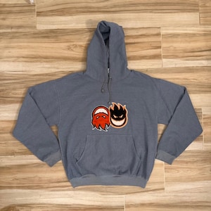 May include: A slate blue hooded sweatshirt featuring a front kangaroo pocket and long sleeves. Centered on the chest is a graphic design showing two stylised flame-like faces, one in red and one in black, both with white teeth and eyes.